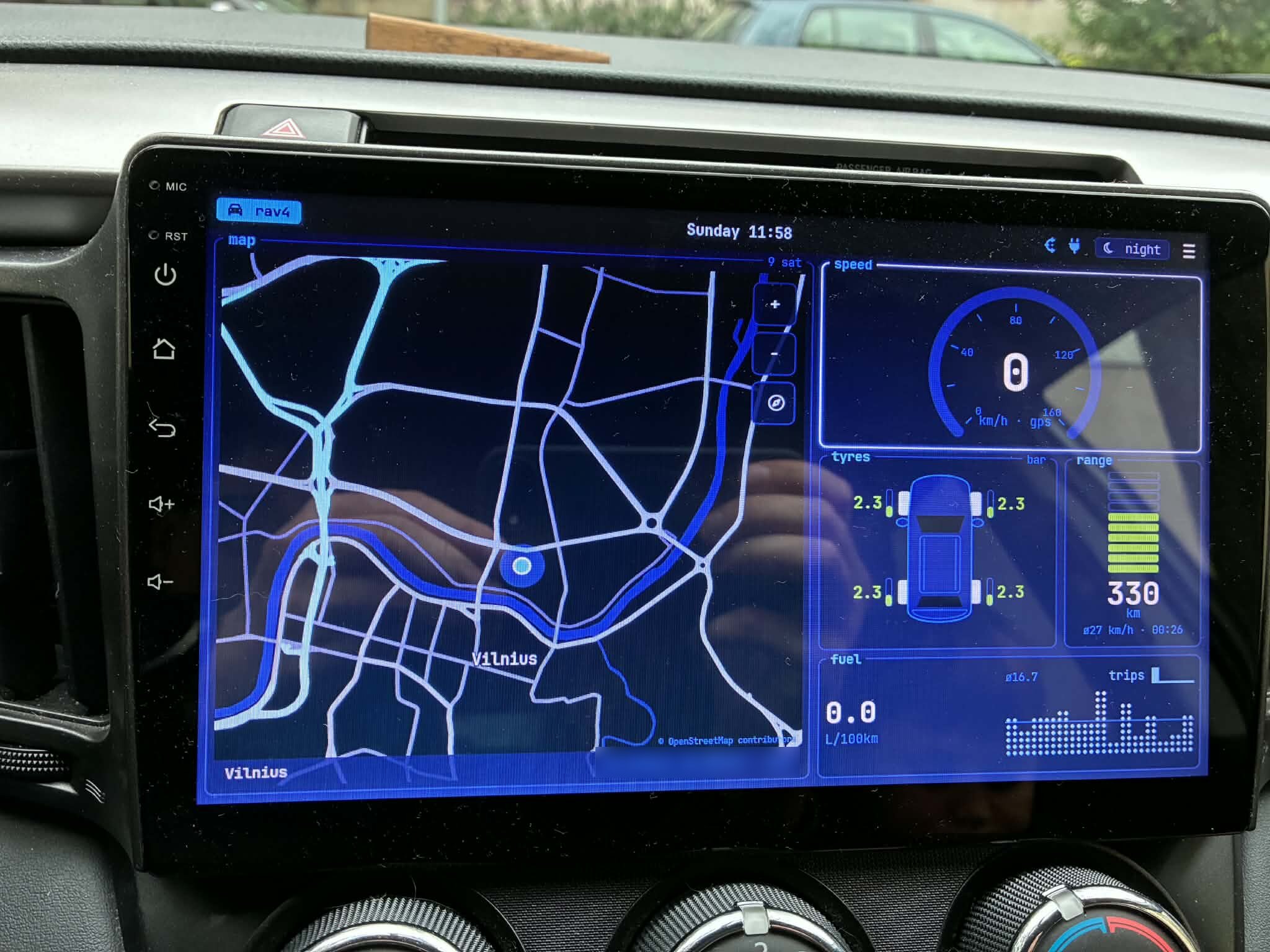 The head unit in the RAV4's dashboard running CarLauncher in night mode: a dark blue offline map of Vilnius on the left, and on the right btop-style panels for a speed gauge, tyre pressures around a top-down car outline (all 2.3 bar), a 330 km range bar, and a dot graph of fuel consumption. A waybar-style top bar shows the car name chip, the clock, status glyphs and a night theme chip.